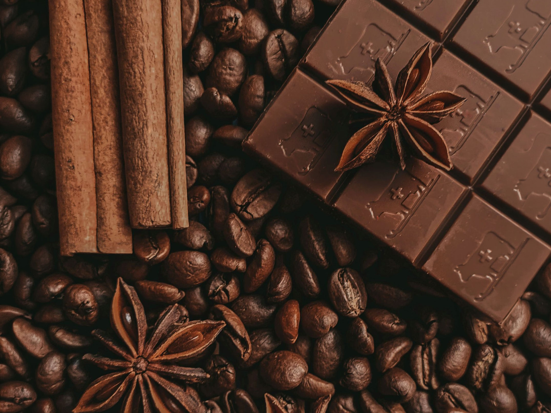 Whole roasted coffee beans spread across a dark surface, with cinnamon sticks, a block of chocolate, and star anise pods